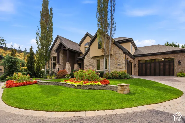 Stone and stucco exterior with a multi-car garage, established landscaping, a paved driveway, and a front lawn with planting beds