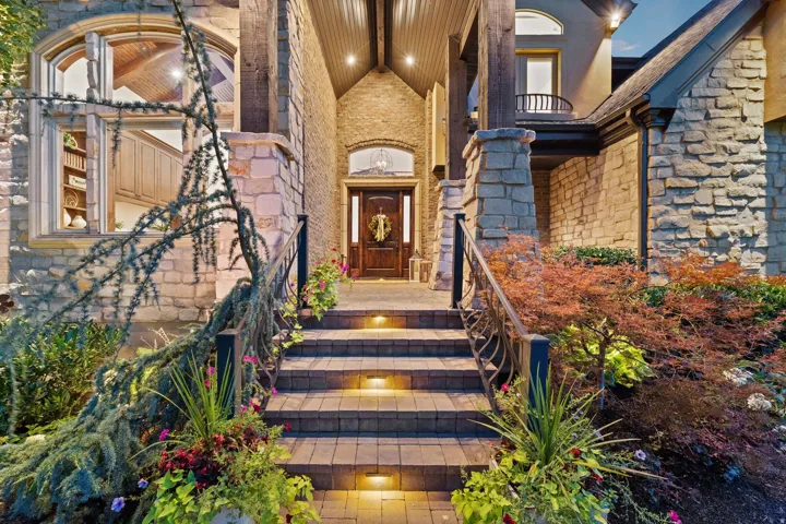 Stone facade exterior featuring an arched wood-finish entry door, paver stone walkway with integrated lighting, and decorative metal railings