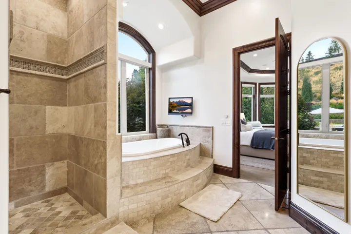 Tiled walk-in shower with decorative mosaic accent, arched window, built-in soaking tub, and stone tile flooring