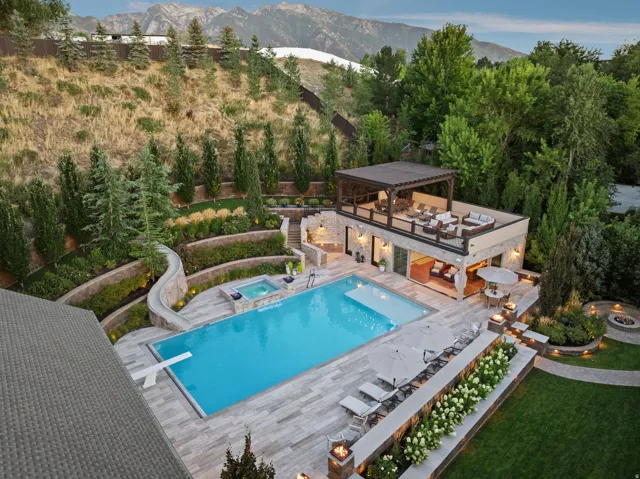 Terraced landscaping with mature trees, expansive swimming pool with diving board, integrated spa, dedicated outdoor structure with an upper-level pergola and lower-level covered patio, and multiple built-in fire features