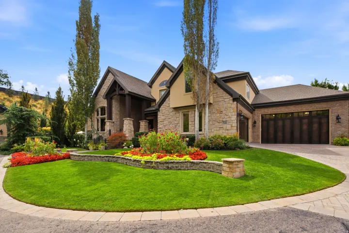 Stone and stucco exterior with a multi-car garage, established landscaping, a paved driveway, and a front lawn with planting beds