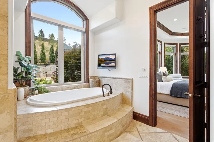 Soaking tub with tiled surround, arched window, wall-mounted television, and wood-framed doorway