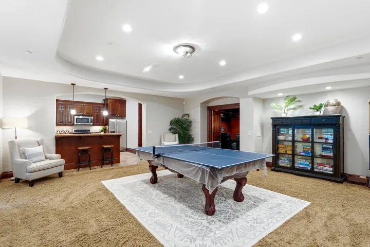 Recessed lighting illuminating a spacious carpeted area with an integrated wet bar featuring wood cabinetry and stainless steel appliances