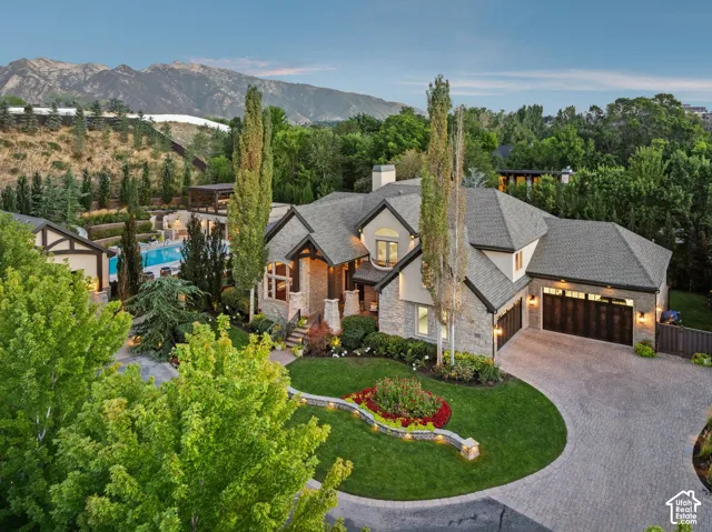 Stone and stucco exterior with multiple gables, three-car garage with wood-finish doors, circular paver driveway, manicured landscaping, and a terraced backyard with a swimming pool