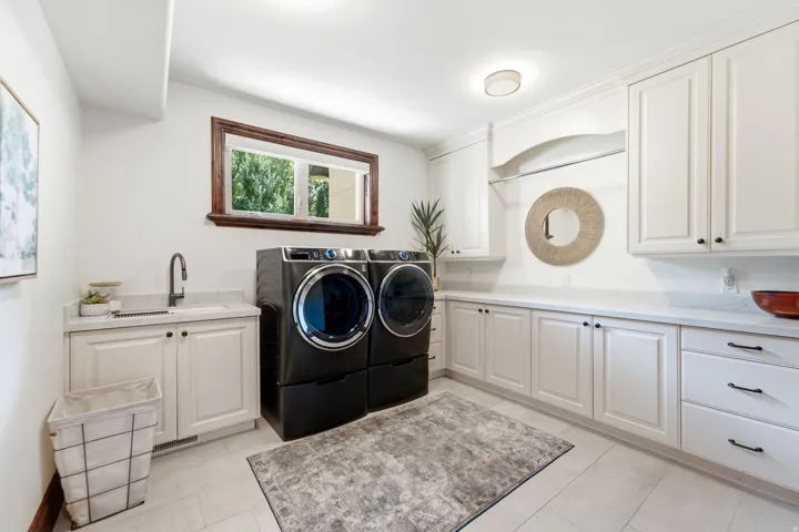 Spacious laundry room with extensive white cabinetry, light-toned countertop surfaces, a utility sink, and wood-framed window