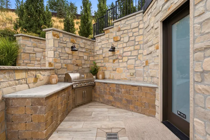 Exterior featuring natural stone walls, a built-in stainless steel grill, light-toned stone countertops, and integrated outdoor lighting fixtures