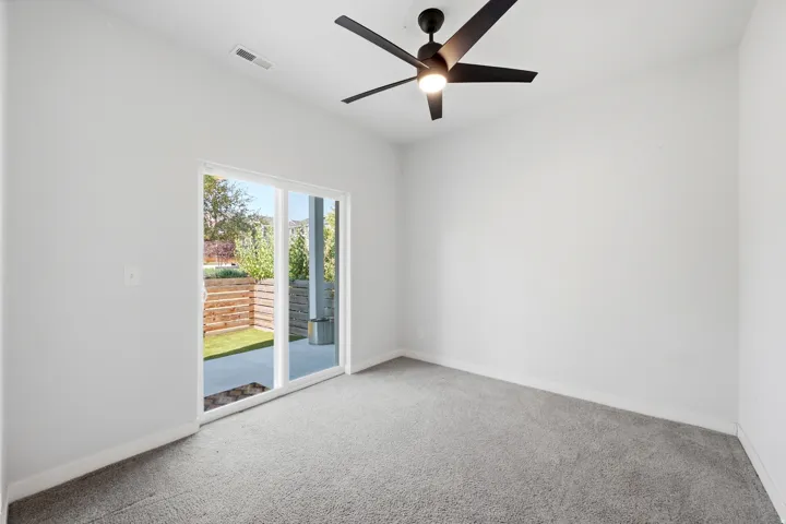 Room featuring a ceiling fan, light gray carpeting, and a sliding glass door opening to an outdoor patio with a wood-slatted retaining wall and artificial turf