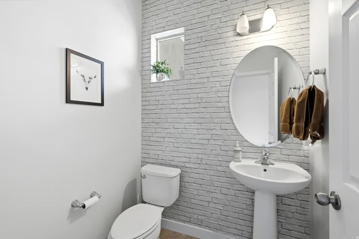 Pedestal sink with chrome faucet, oval mirror, wall-mounted light fixture, and textured brick-patterned wall