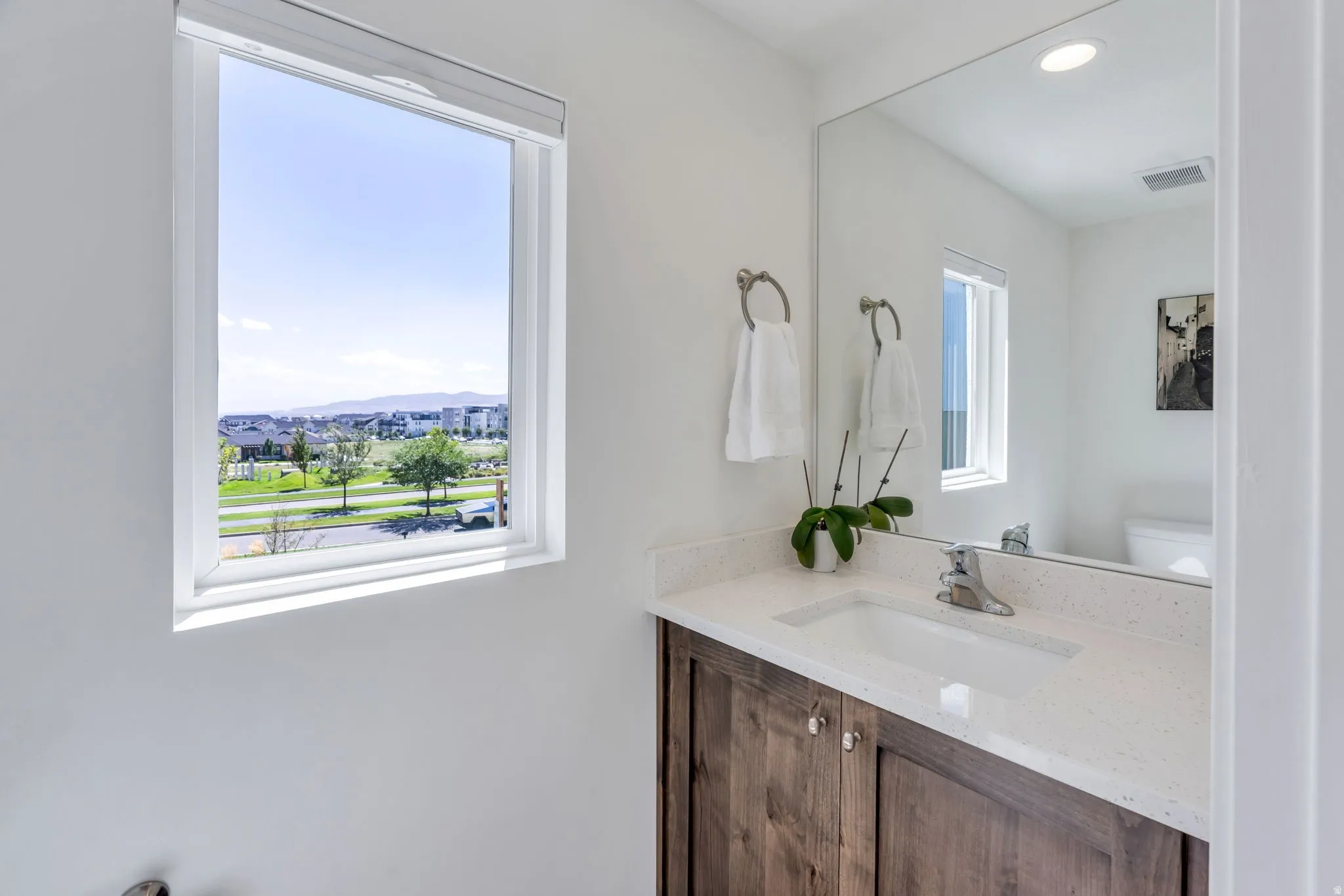 White walls with a single window, a wood-finish vanity with a white countertop and integrated sink, and a large wall-mounted mirror