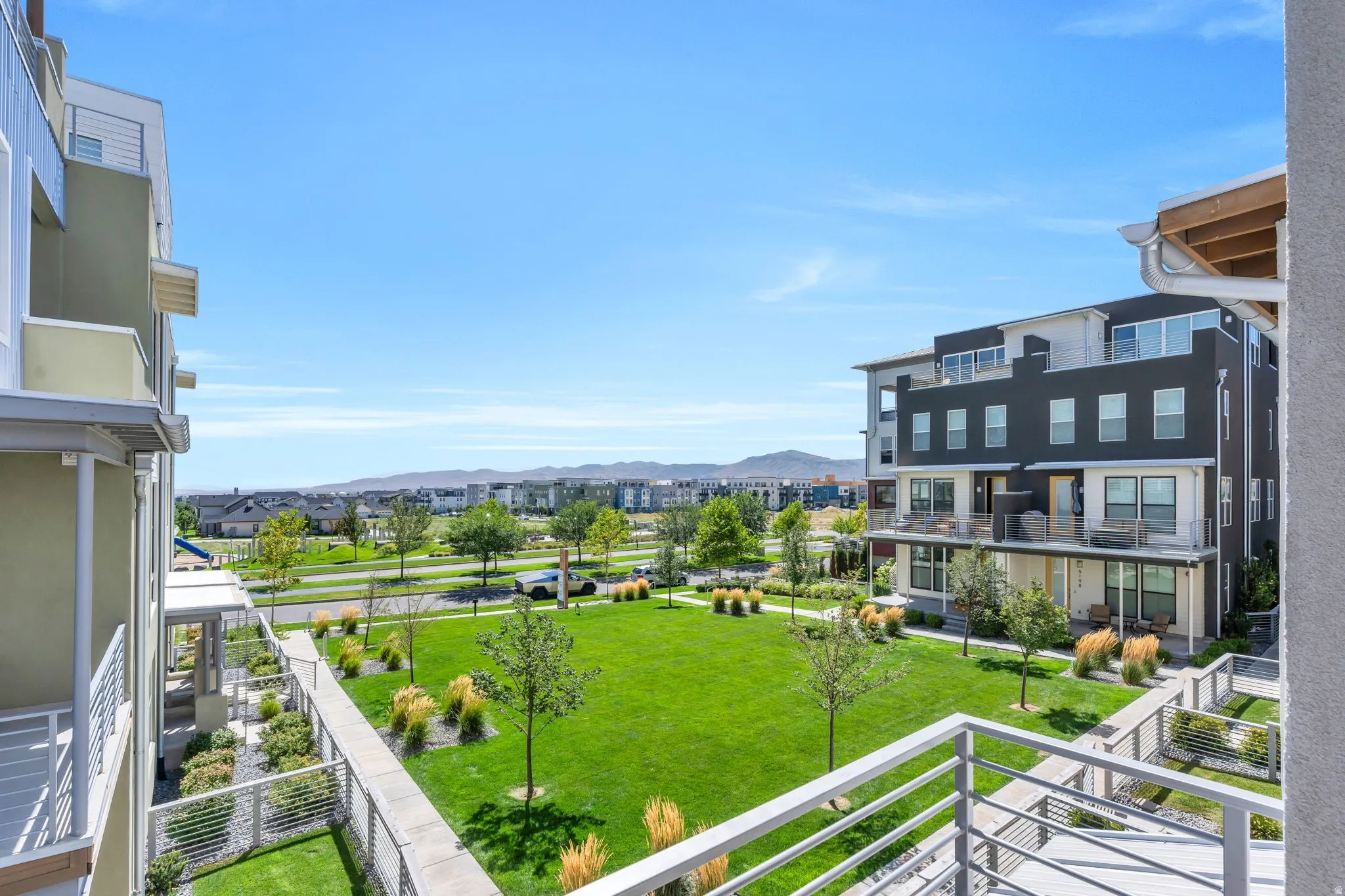 Balcony with metal railings overlooking a landscaped common area with expansive green lawn and mature plantings, surrounded by contemporary multi-story structures
