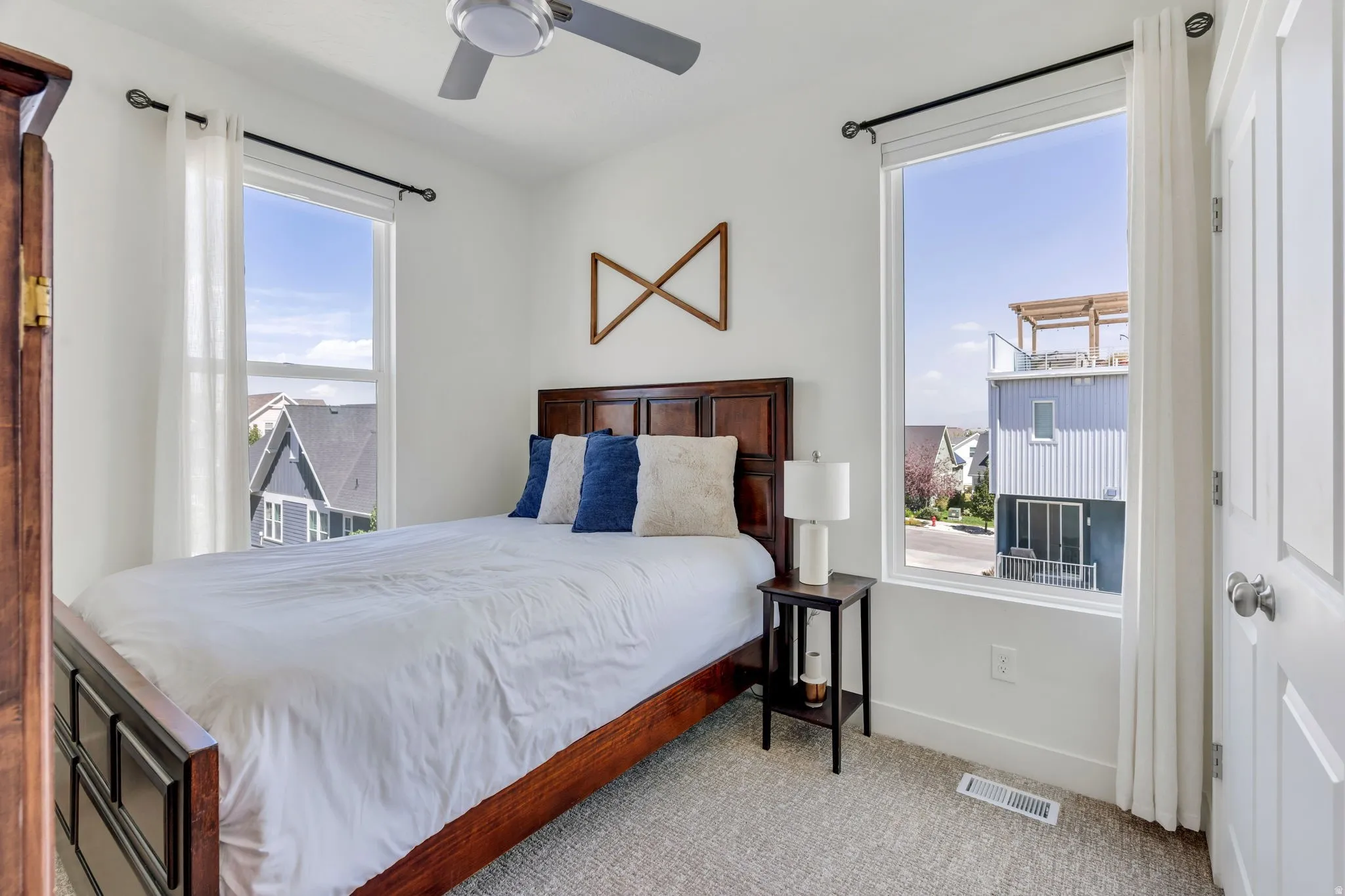 White walls, two large windows, a ceiling fan, a dark wood-finish bed frame, and light-toned carpet