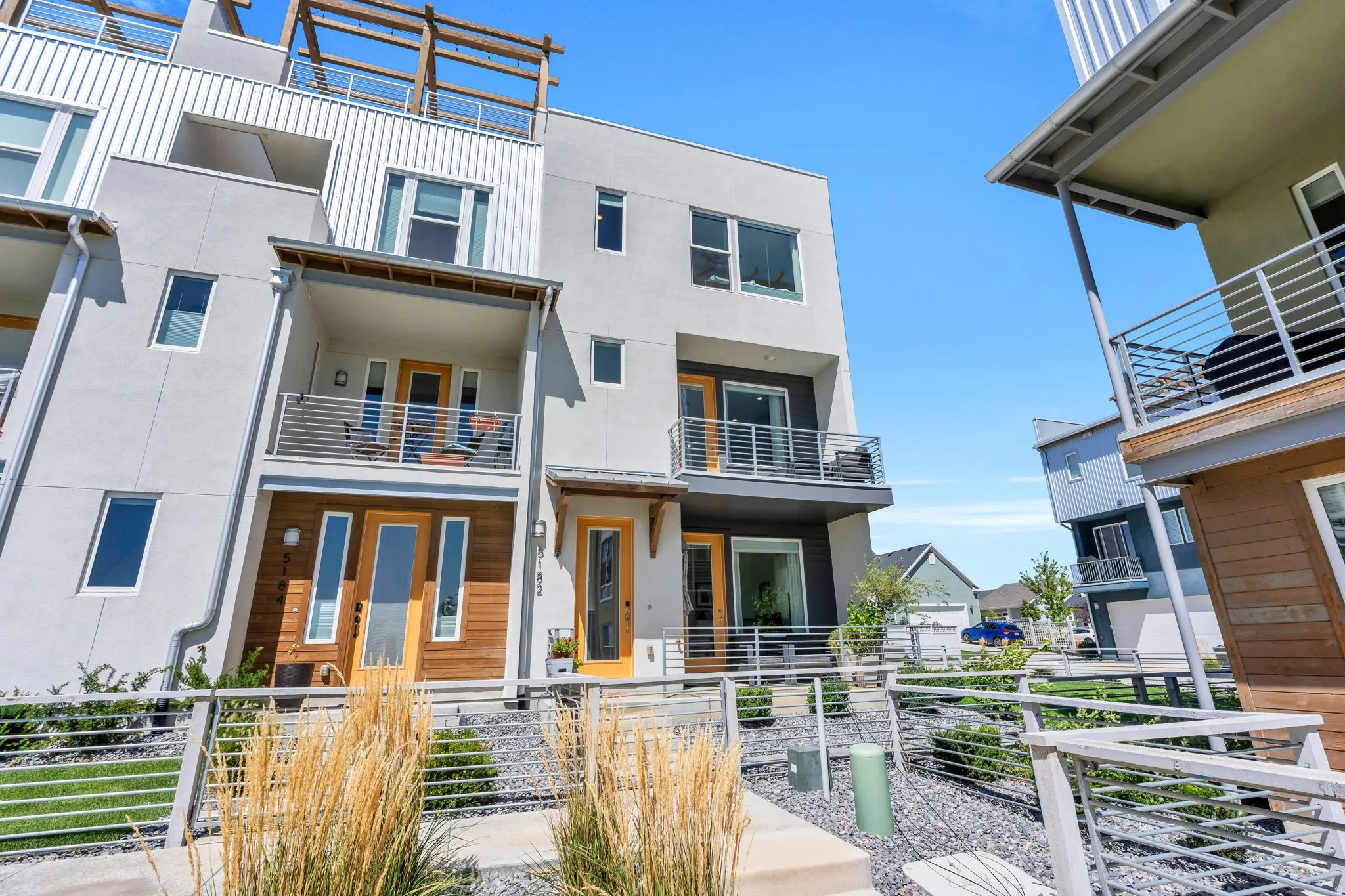 Contemporary multi-story exterior with stucco and wood paneling, featuring multiple balconies with metal railings, a rooftop deck with a wood pergola, and concrete walkways