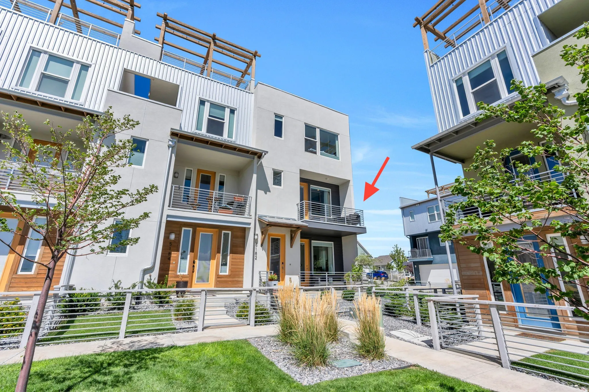 Modern townhouse exterior featuring a private balcony with cable railings, wood-finish accent paneling, a glass-paneled entry door, and a low-maintenance landscaped walkway