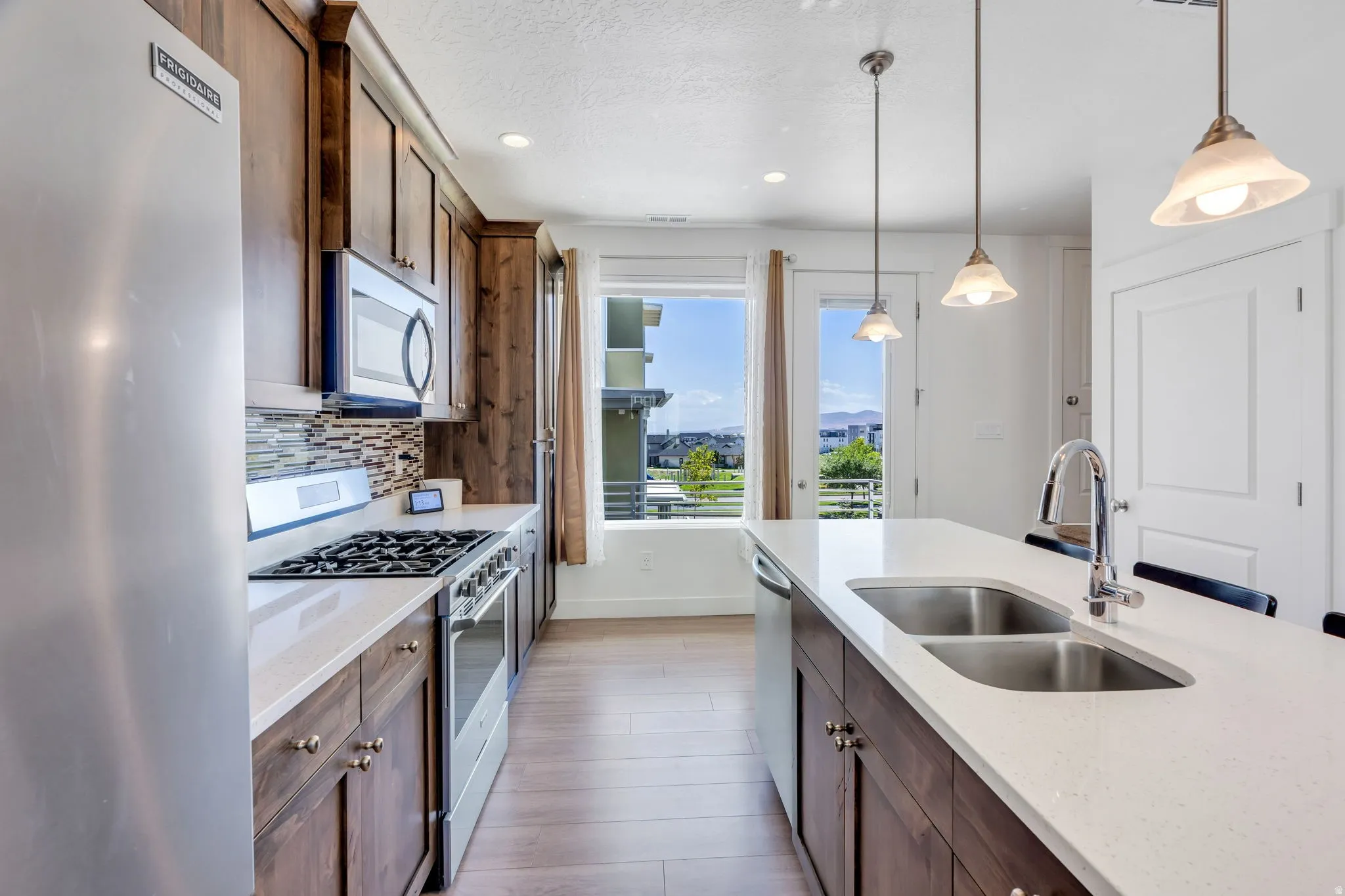 Kitchen featuring wood-finish cabinetry, stainless steel appliances, light-toned countertops, a mosaic tile backsplash, and an island with a double basin sink