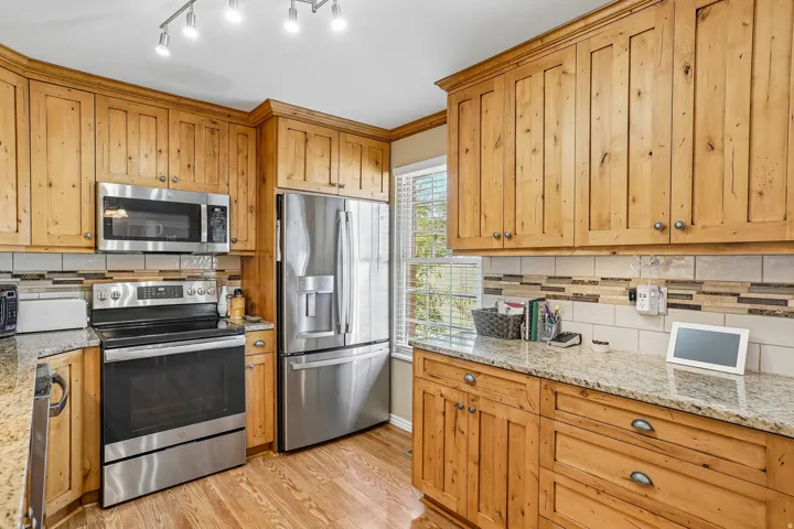 Stainless steel appliances, natural wood cabinetry, light-toned granite countertops, wood-finish flooring, and a tiled backsplash