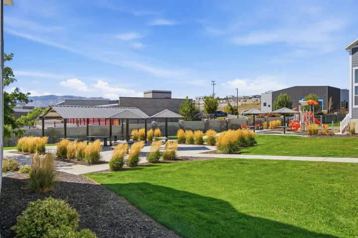 Expansive green space located to the side of the townhome, featuring a paved walkway, covered picnic areas, a children's play structure, and landscaped planting beds