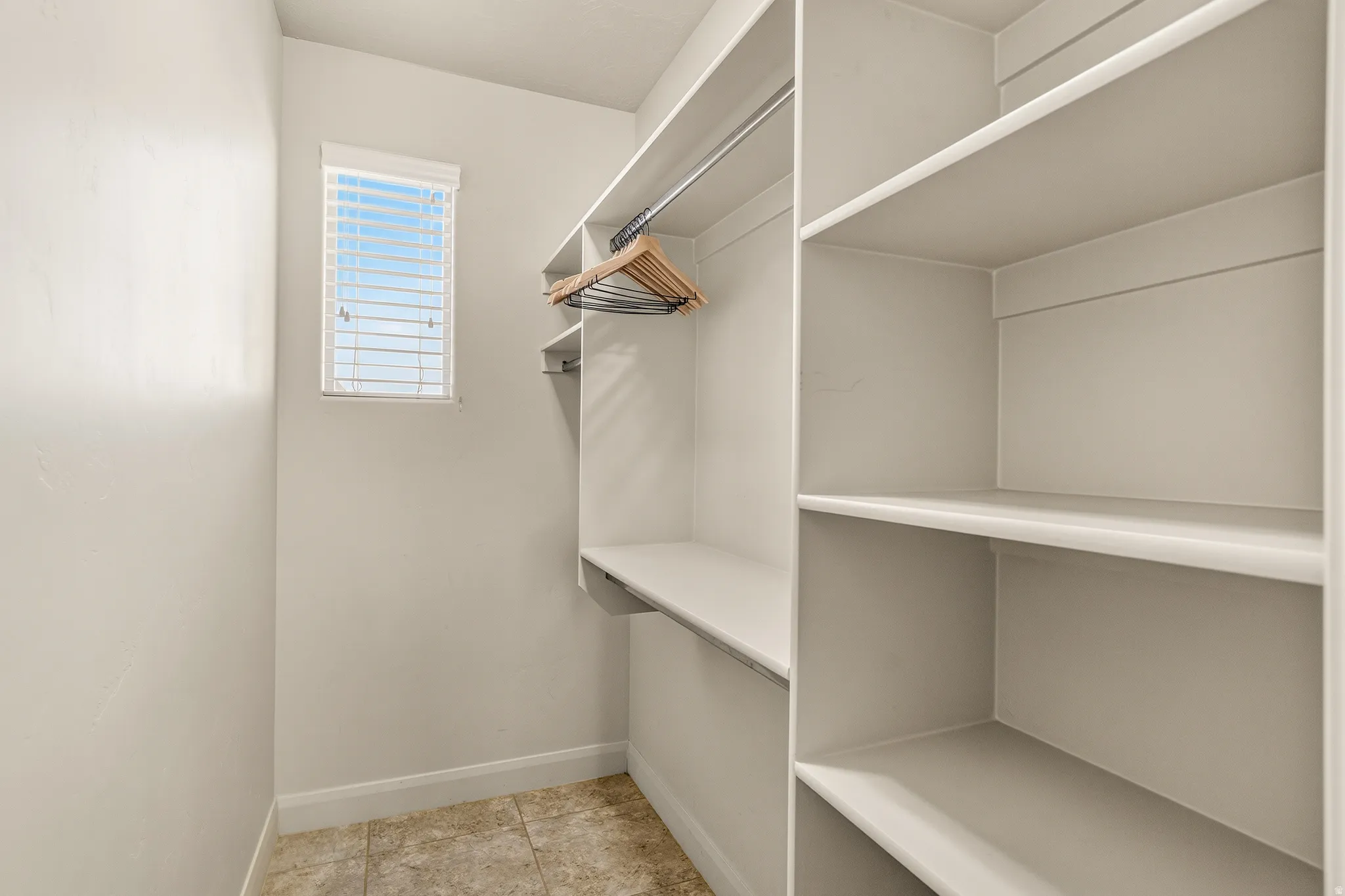 Built-in shelving, hanging rods, window with blinds, and tile flooring