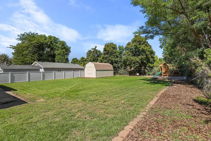 Expansive grassy yard with a white privacy fence, mature trees, a storage shed, and a mulched garden bed with brick edging