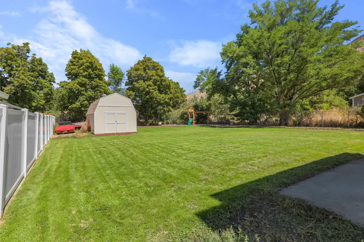 Expansive grassy yard featuring a storage shed, mature trees, and a concrete patio