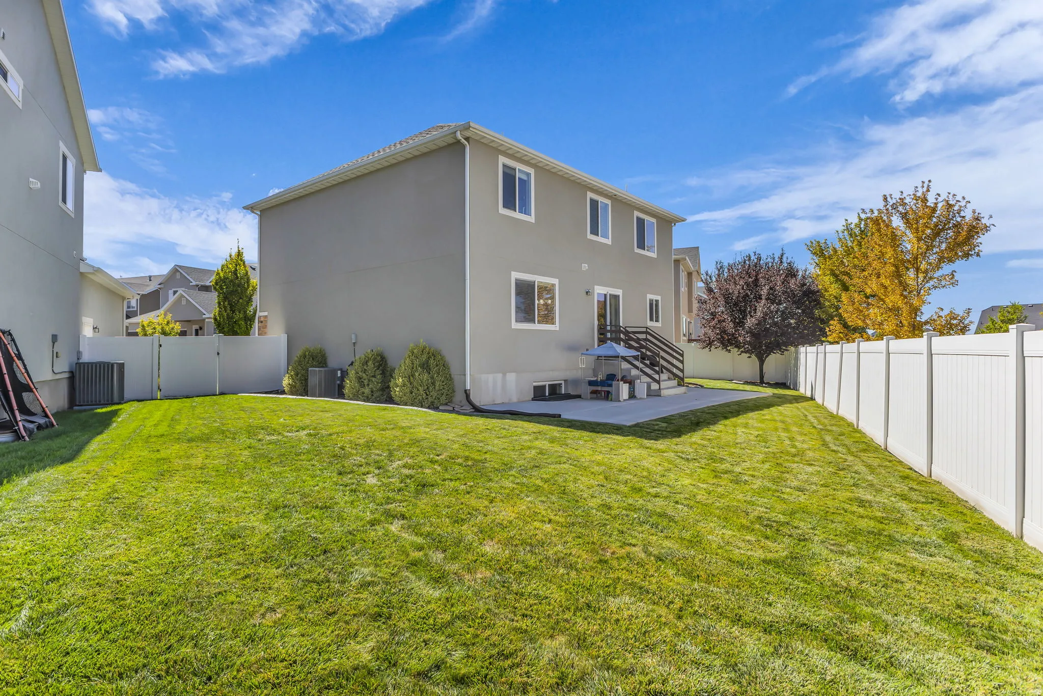 Expansive green lawn, white privacy fencing, concrete patio, exterior stairs with dark railings, and a stucco exterior finish