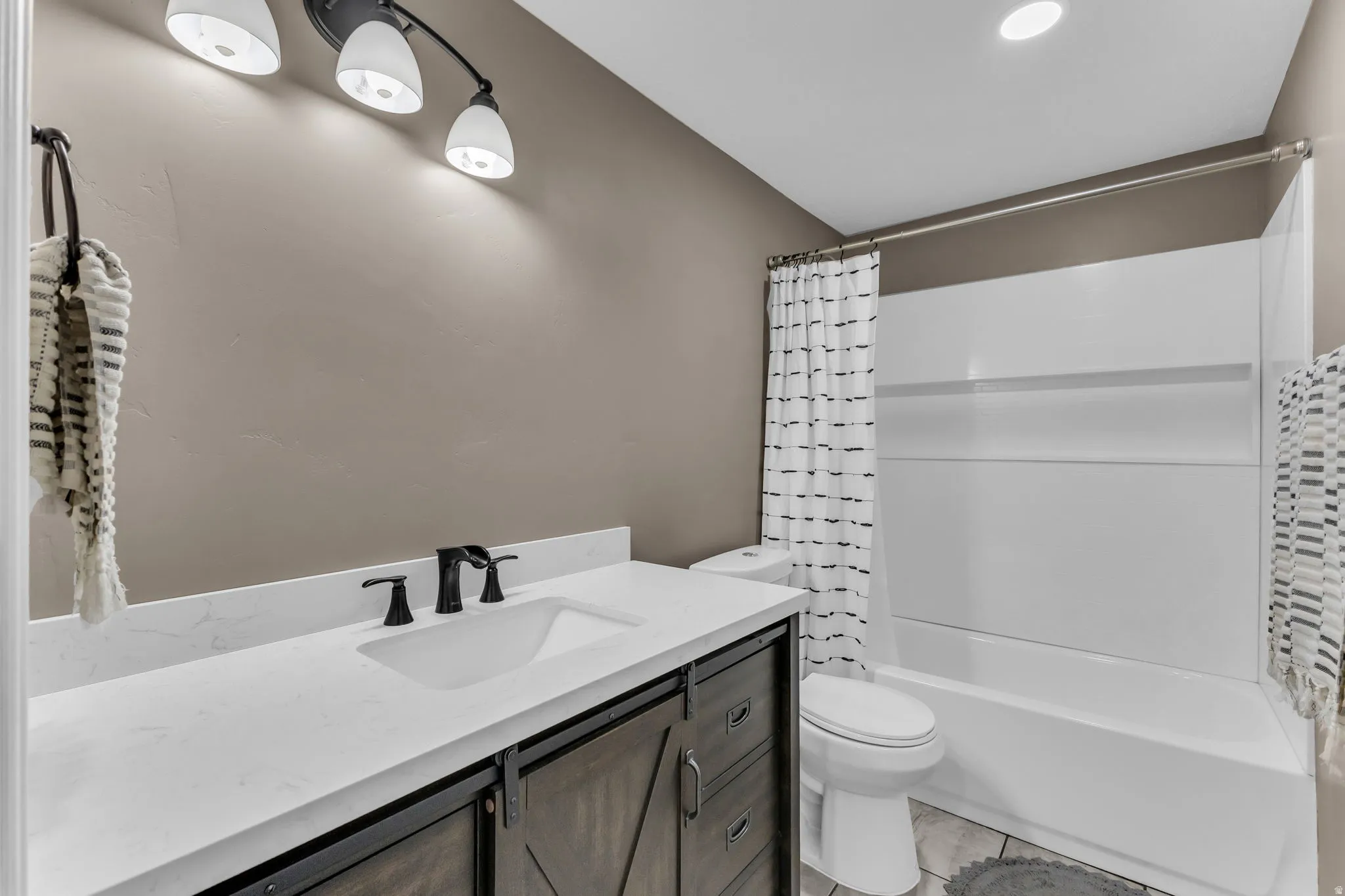 White countertop with integrated sink, black matte faucet, wood-finish vanity with barn door hardware, white bathtub with shower, and tile flooring