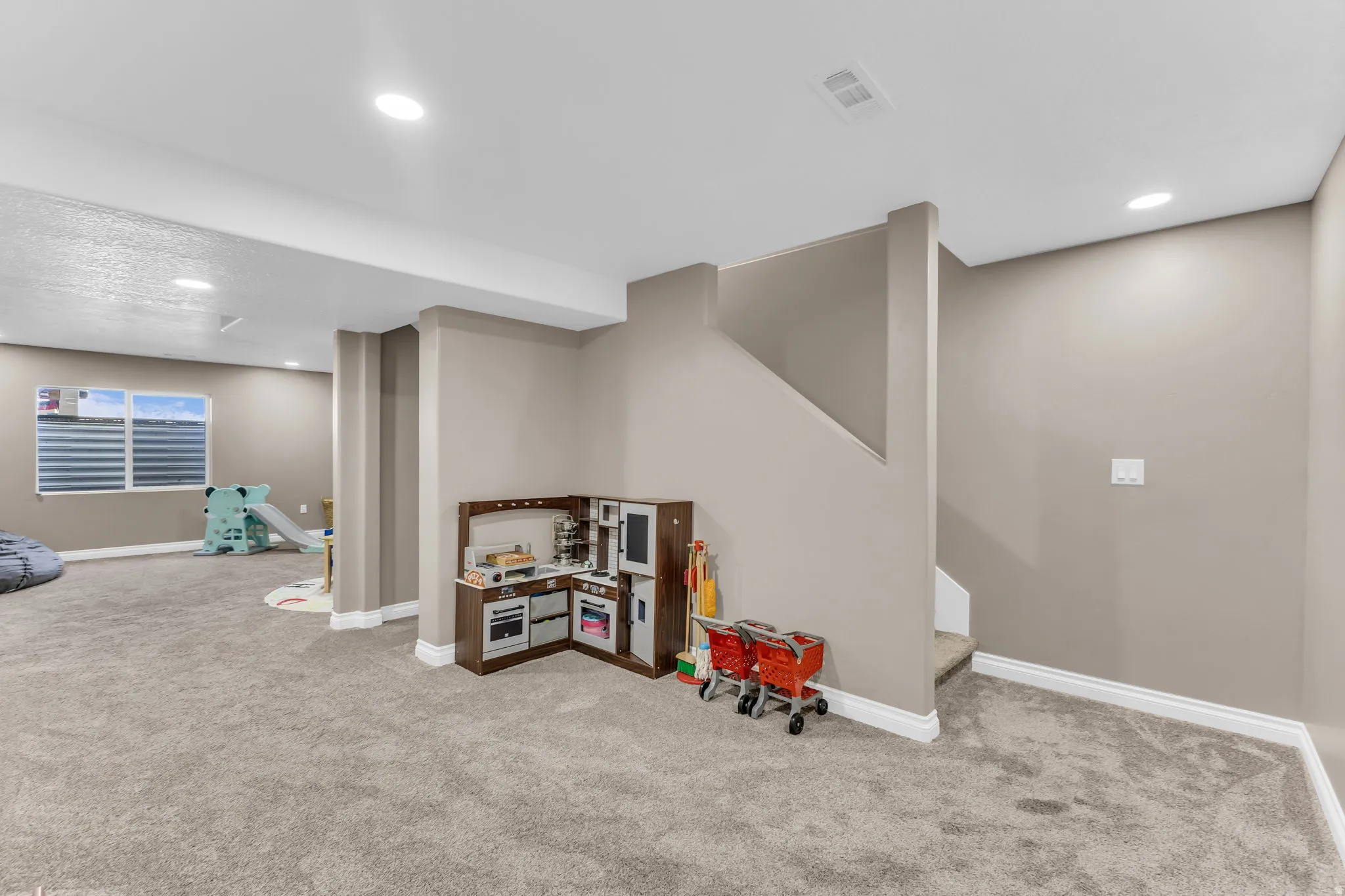 Carpeted room with recessed lighting, neutral wall tones, a visible window, and a staircase