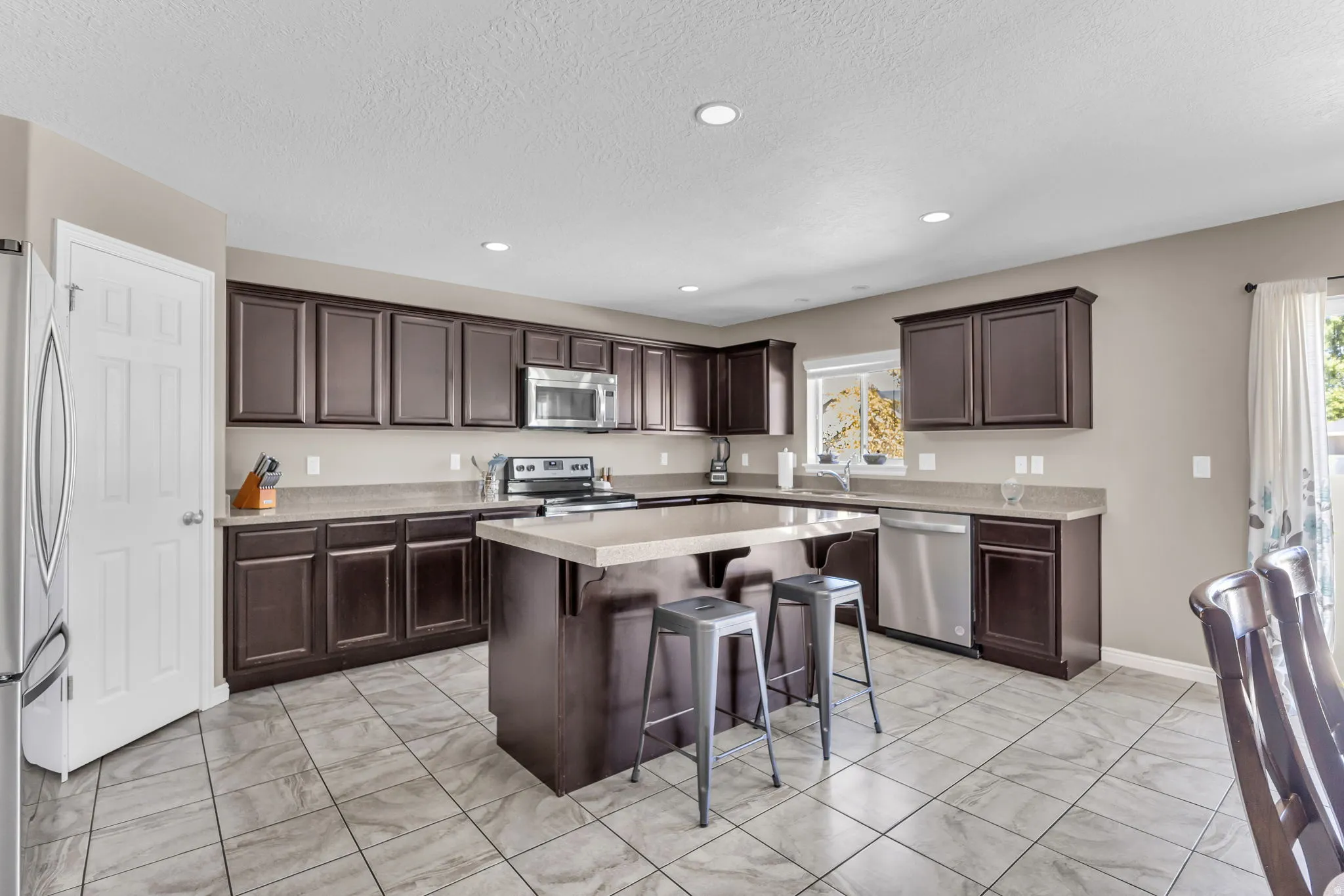 Kitchen featuring dark wood-finish cabinetry, light-toned countertops, a central island, stainless steel appliances, and tiled flooring