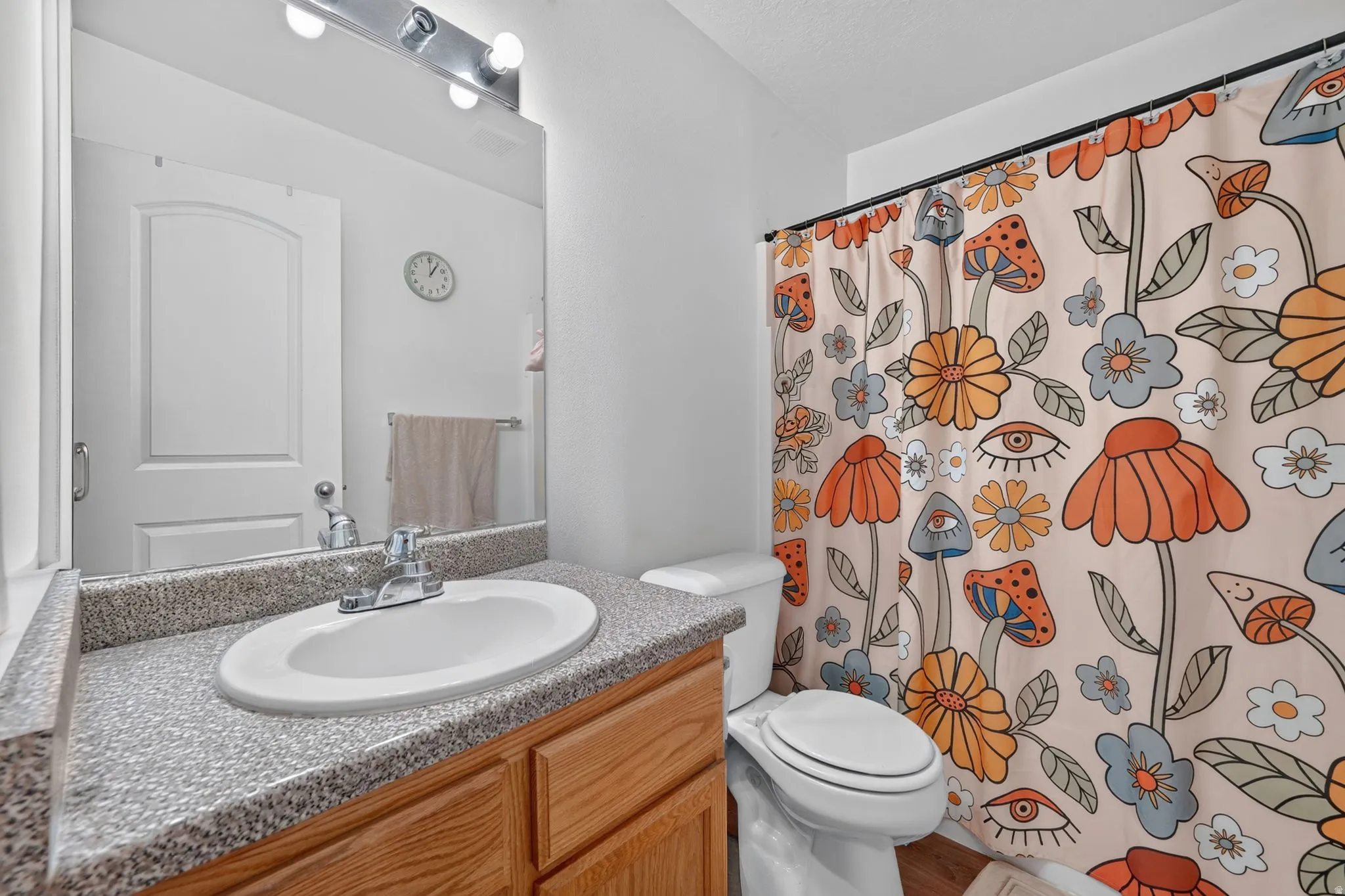 Spa-inspired bathroom featuring a wood-finish vanity with dual-basin granite countertop, recessed lighting above the mirror, and a decorative shower curtain with whimsical botanical and floral motifs