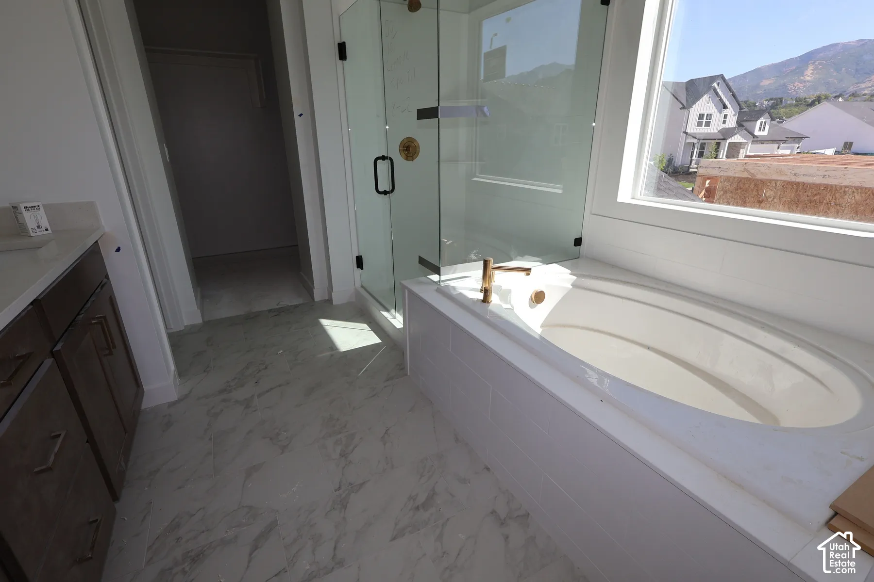 Soaking tub with brass fixtures, glass-enclosed shower, wood-finish vanity with countertop, and marble-look tile flooring