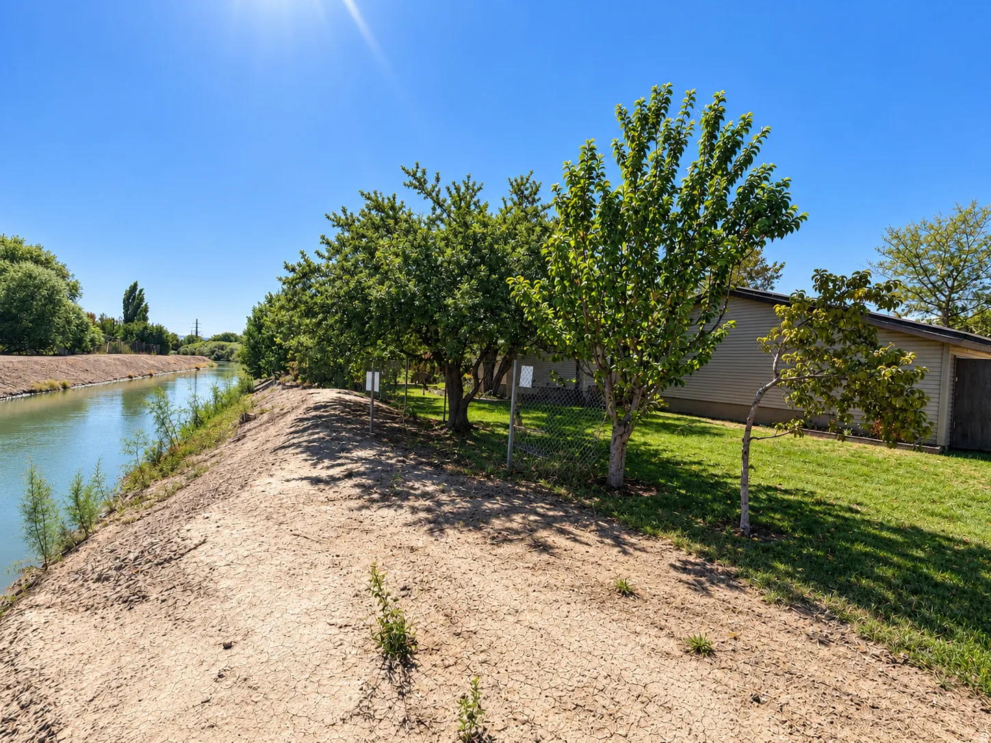 Canal frontage with a natural bank, mature trees, and a grass lawn. Chain link fence along the back