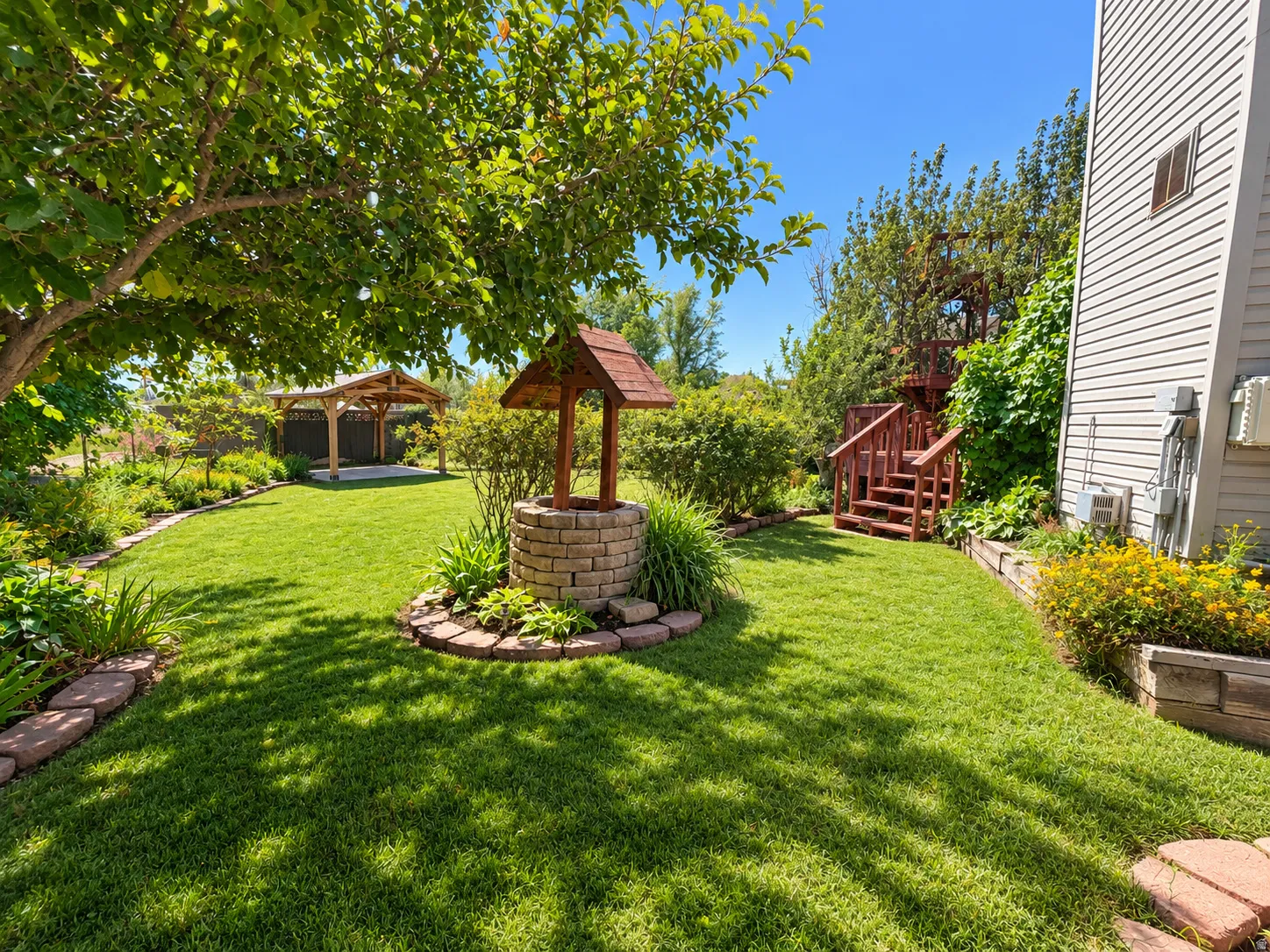 Expansive green lawn with a decorative brick wishing well, mature trees, a wooden gazebo structure, and a multi-level wooden staircase