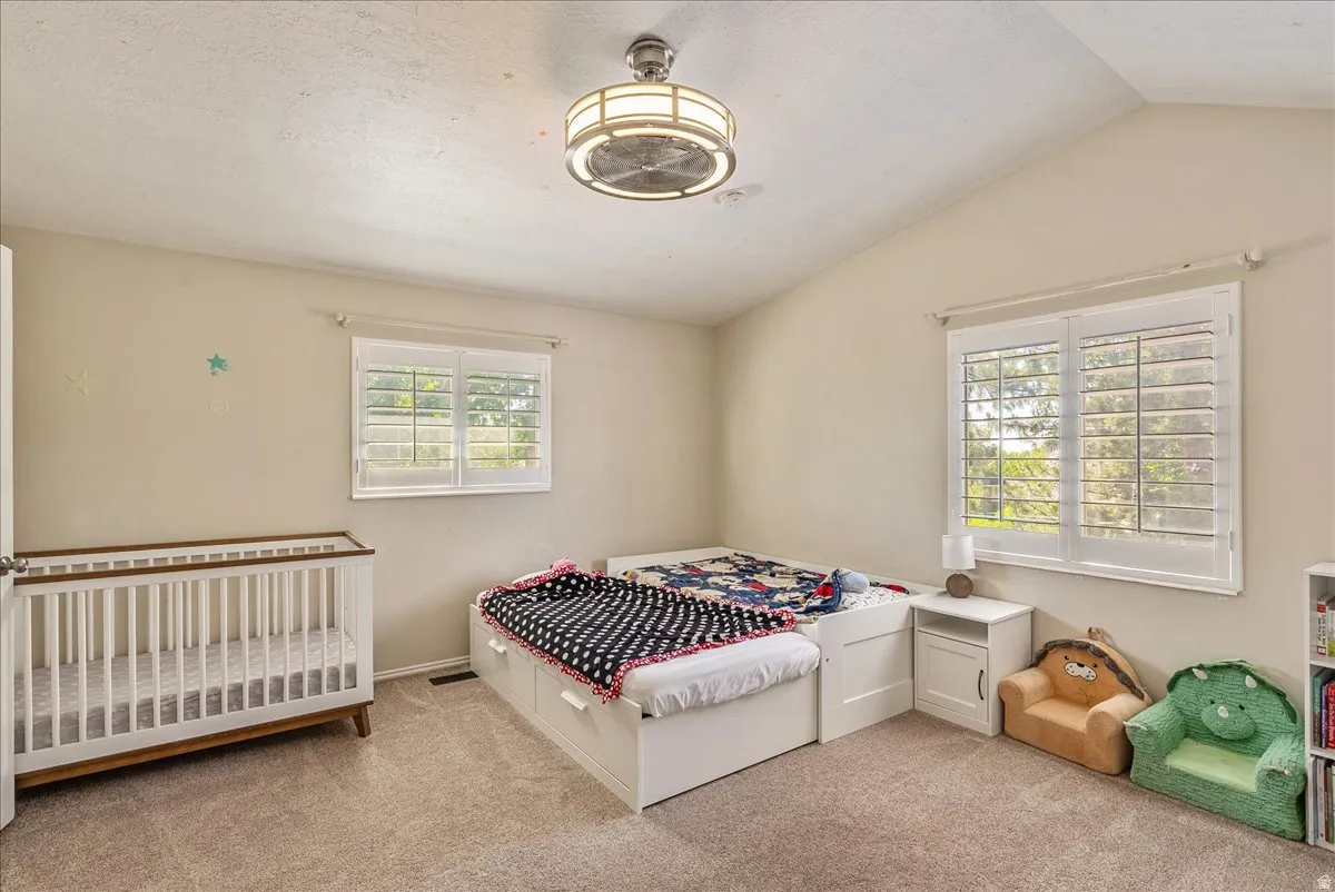 Vaulted ceiling, two windows with plantation shutters, ceiling fan with integrated lighting, and carpeted flooring