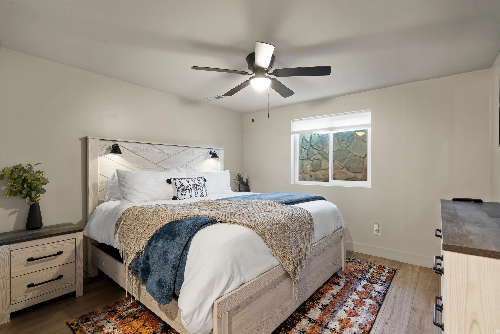 Wood-finish flooring, ceiling fan with integrated lighting, shiplap-style headboard with sconce lighting, and a horizontal window