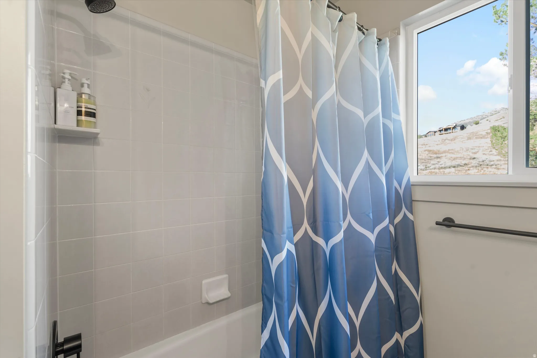 Tub and shower combination featuring light gray tile surround, matte black shower head, and a window providing natural illumination