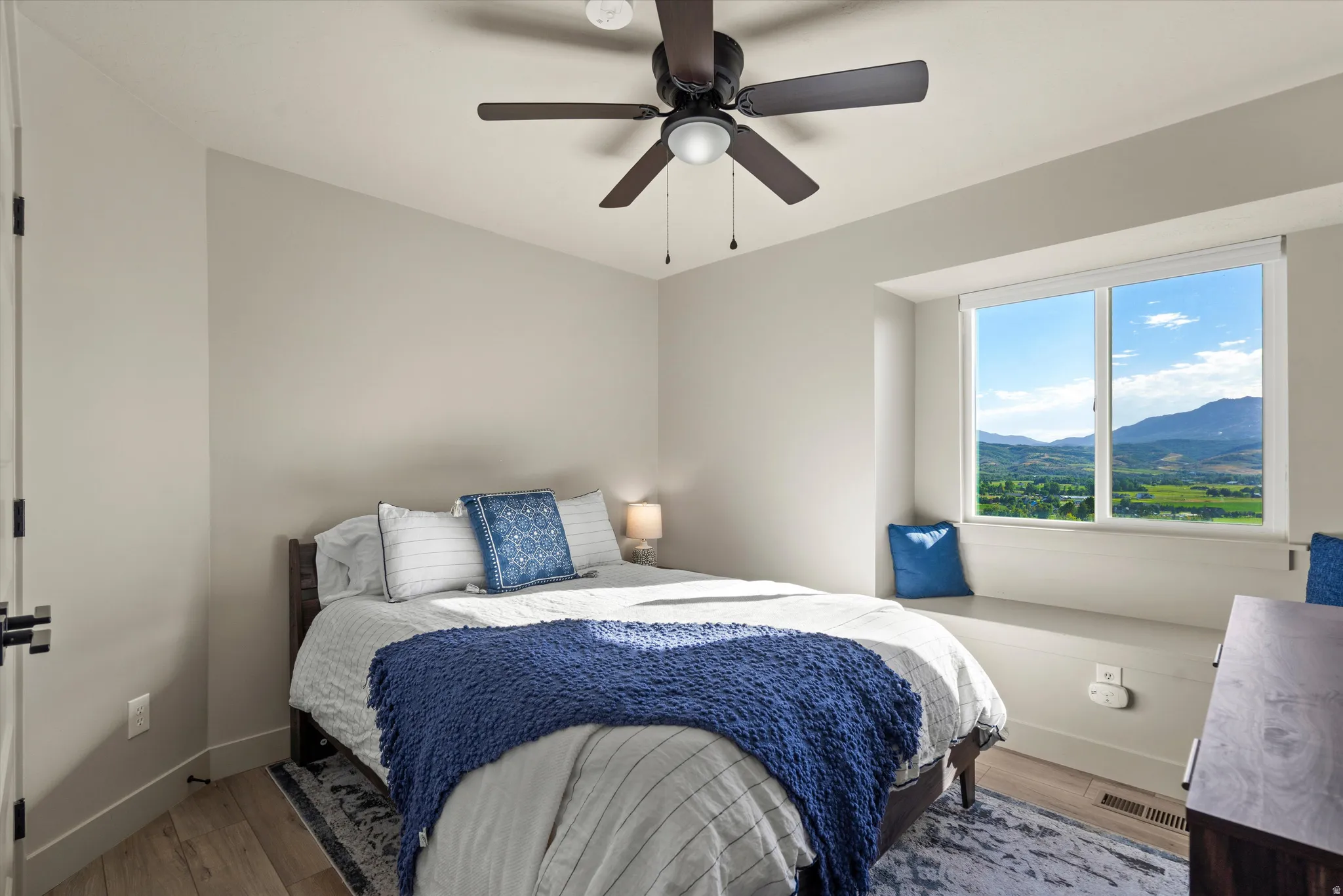 Light wall color, window seat, mountain scenery, wood-finish flooring, and ceiling fan
