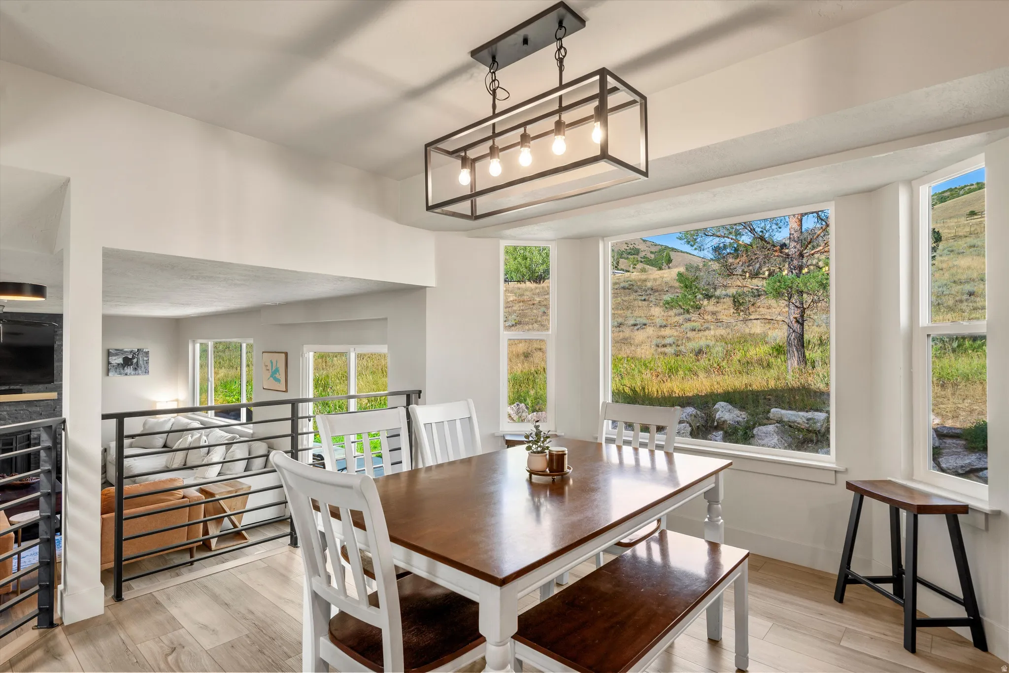 Open-concept dining area featuring wood-finish flooring, a contemporary linear chandelier, expansive bay windows, and a metal railing overlooking a lower level