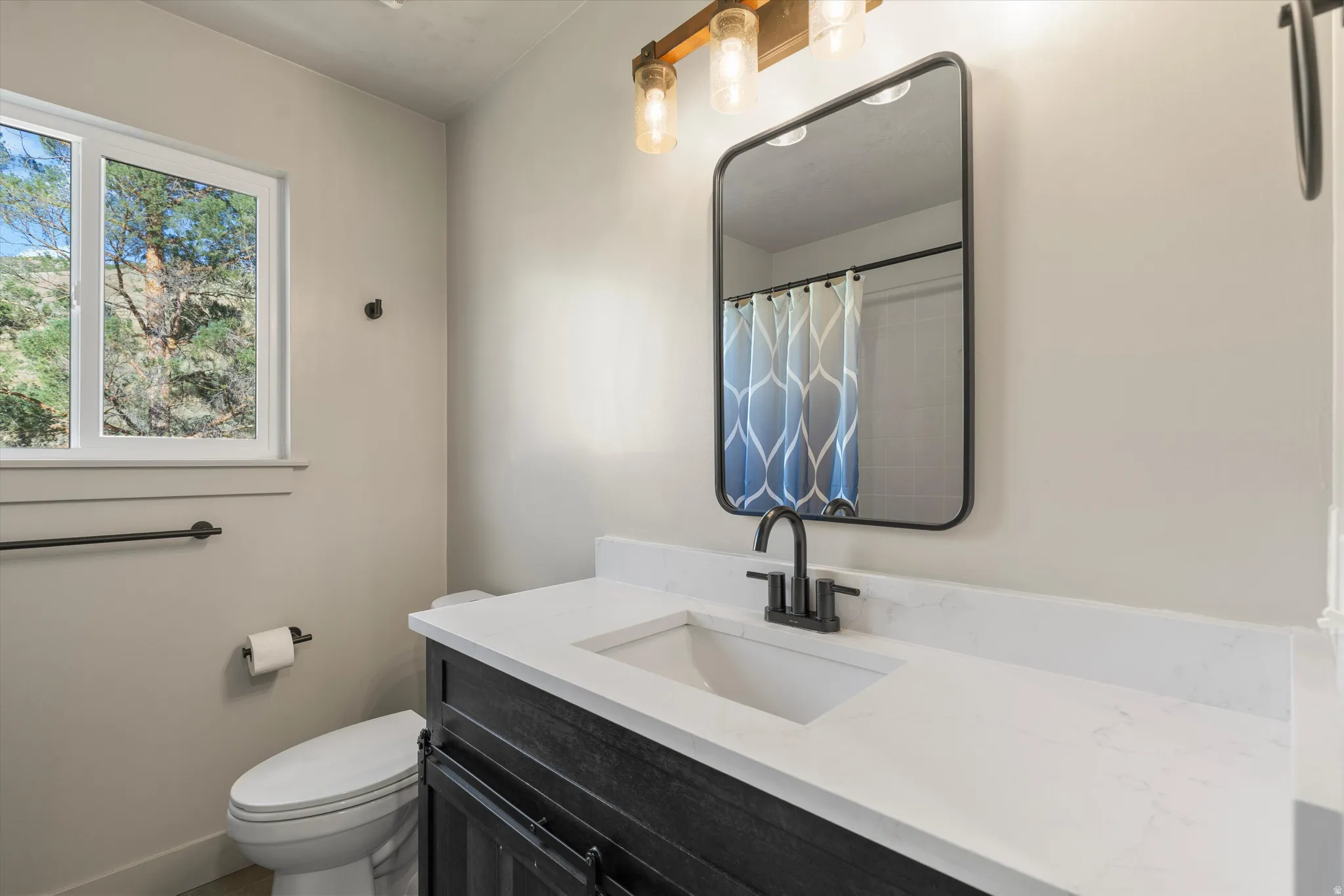 White-framed window, vanity with a white countertop, undermount sink, and black faucet, rectangular wall mirror with a black frame, and a three-light vanity fixture