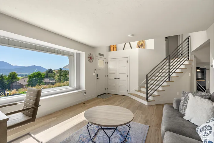 Expansive picture window, light wood-finish flooring, contemporary open-tread staircase with horizontal metal railing, white paneled doors, and architectural high ledges