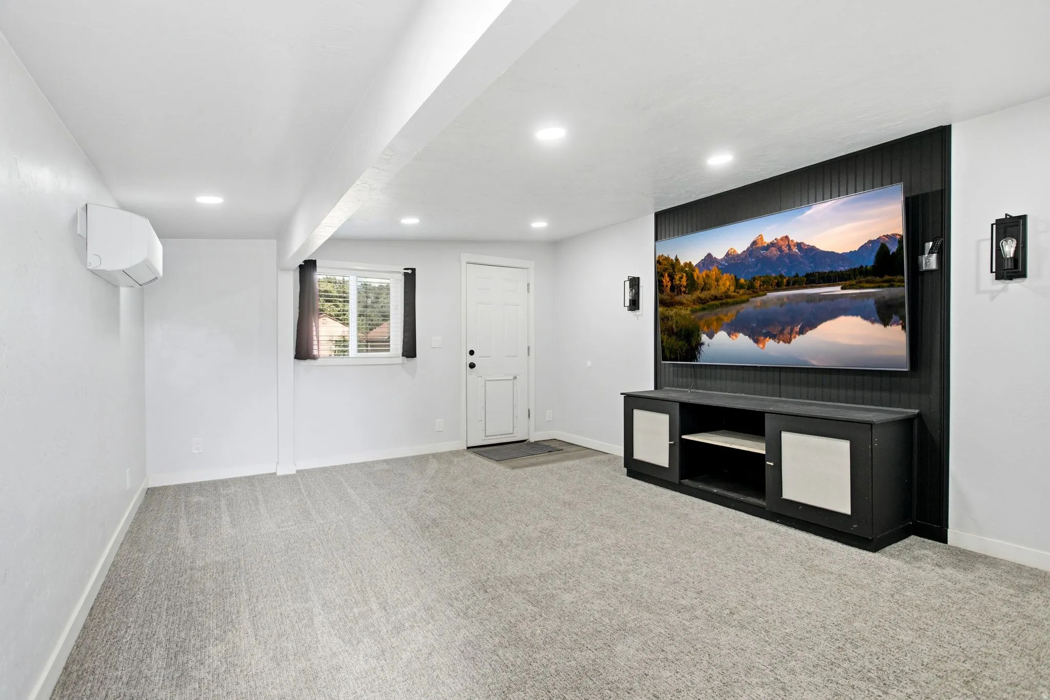 Recessed lighting illuminates the carpeted room, featuring a built-in media console with storage, a wall-mounted mini-split AC unit, and a window with blinds