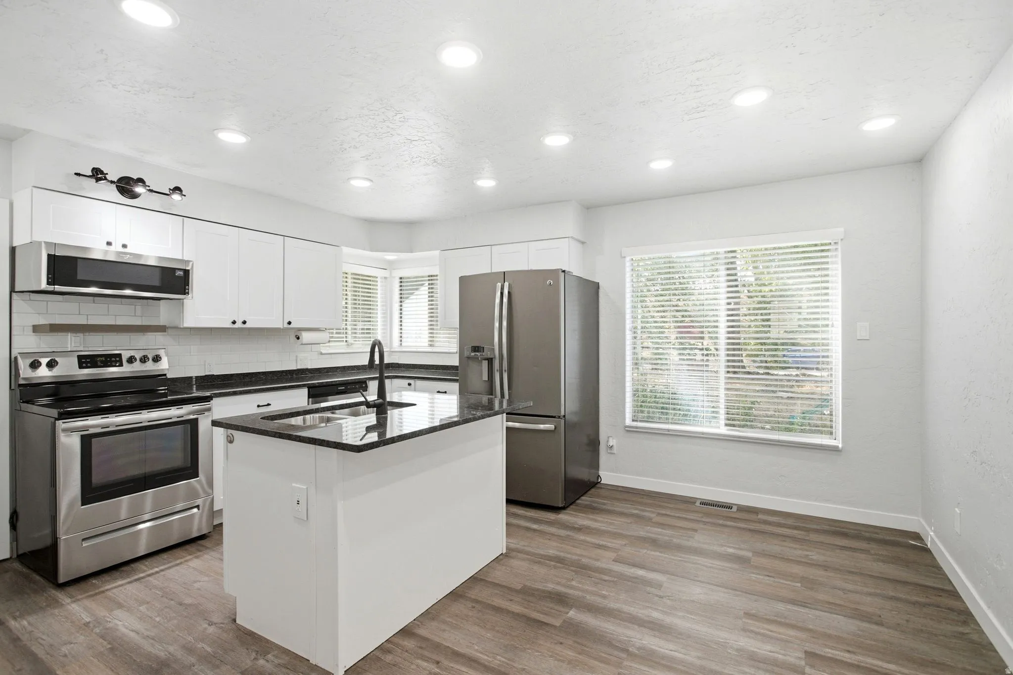 Modern kitchen featuring stainless steel appliances, white cabinetry with black countertops, a central island with seating space, wood-finish flooring, recessed ceiling lighting, and expansive windows providing natural light throughout the space