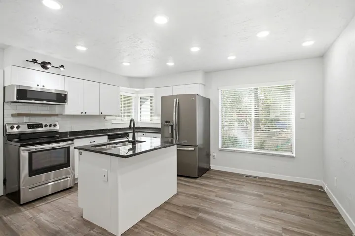 Modern kitchen featuring stainless steel appliances, white cabinetry with black countertops, a central island with seating space, wood-finish flooring, recessed ceiling lighting, and expansive windows providing natural light throughout the space