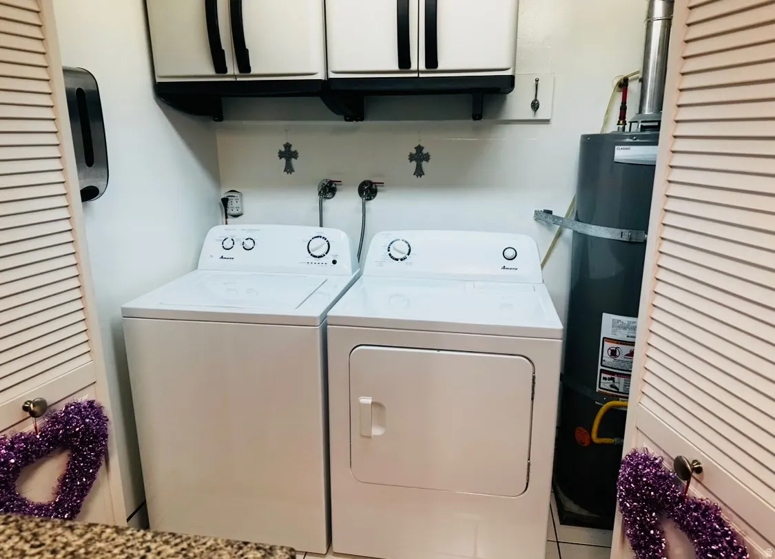 Dedicated laundry room featuring side-by-side washer and dryer with white finish, overhead cabinetry with black hardware, decorative wall crosses, and tile flooring throughout the space