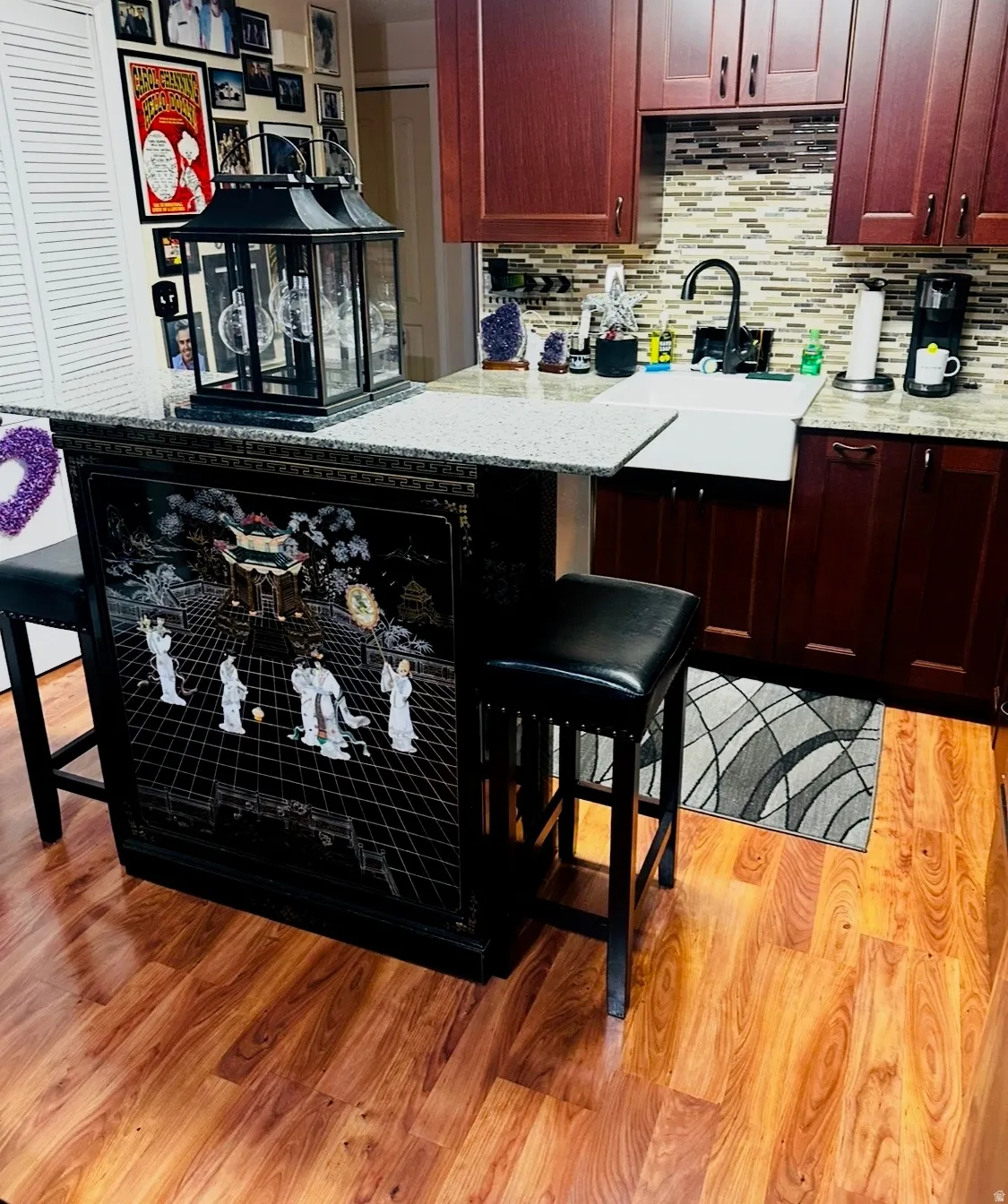 Wood-finish flooring, dark wood-tone cabinetry, tiled backsplash, white farmhouse-style sink, and light-toned countertop