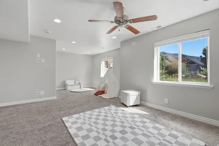 Recessed ceiling lighting, ceiling fan with wood-finish blades, light gray carpeting, and a bright window with white trim
