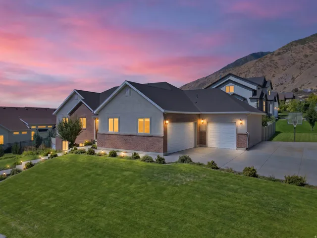 Stucco and brick exterior with a dark shingle roof, attached two-car garage, concrete driveway, and an expansive lawn with landscape lighting