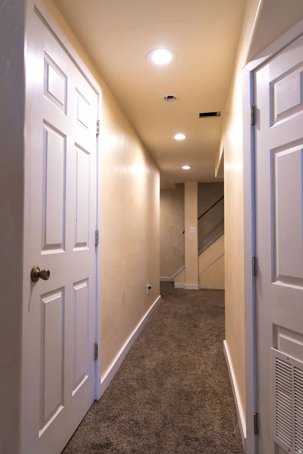 Recessed lighting illuminates a hallway with paneled interior doors and brown carpeting