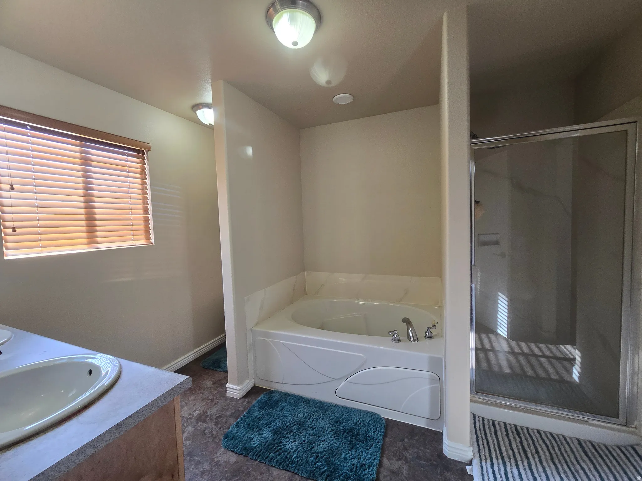 Soaking tub with integrated jets, glass-enclosed shower, dual vanity with undermount sink, window with blinds, and flush-mount lighting fixtures