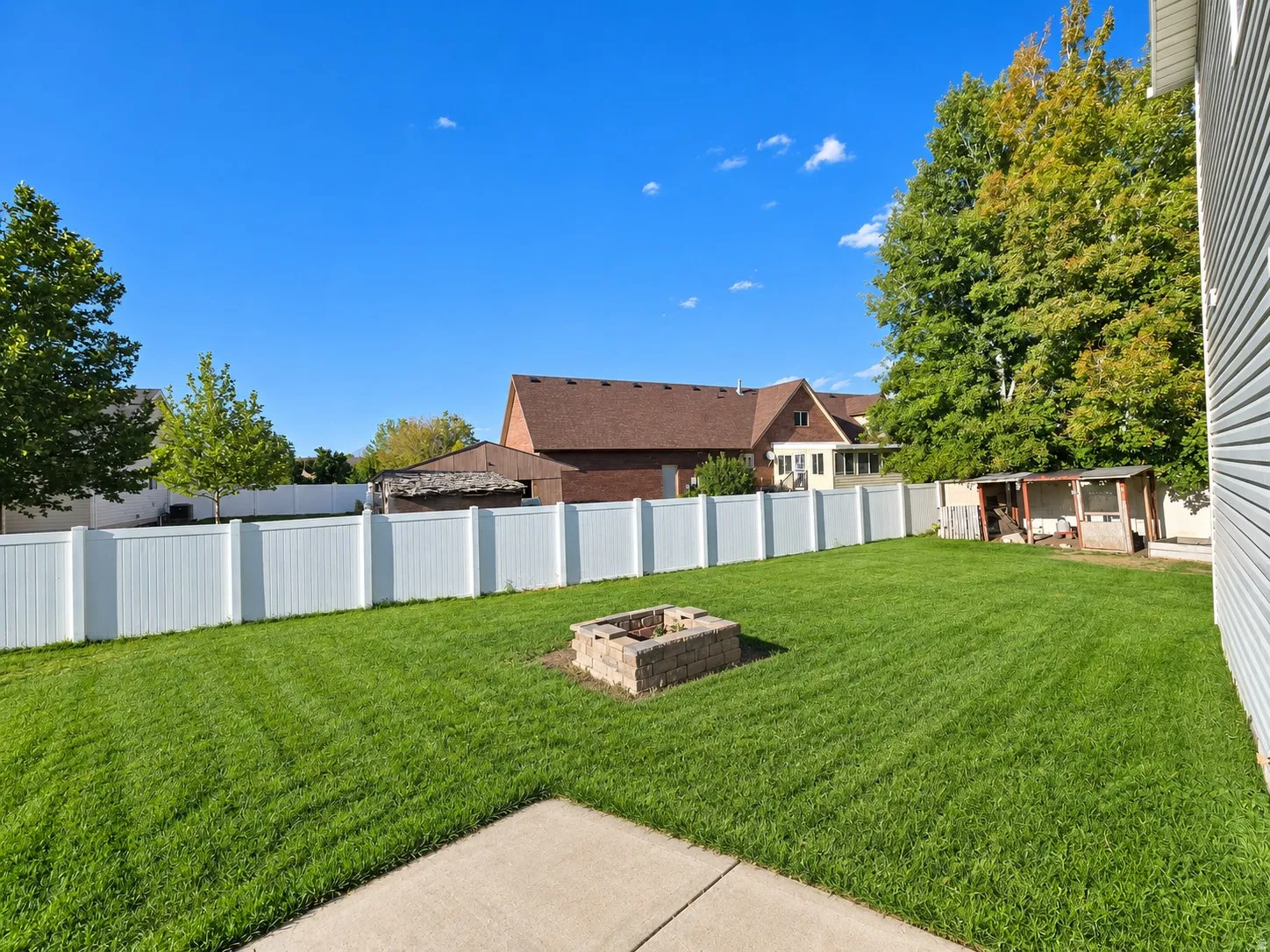 Expansive green lawn featuring a stone fire pit, bordered by a white privacy fence, and a concrete patio section