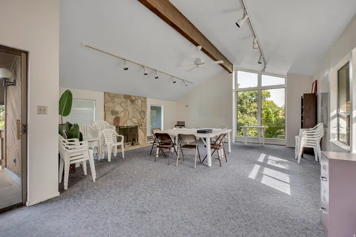 Vaulted ceiling with exposed wood beams, stone-clad fireplace, track lighting, ceiling fan, and large picture windows