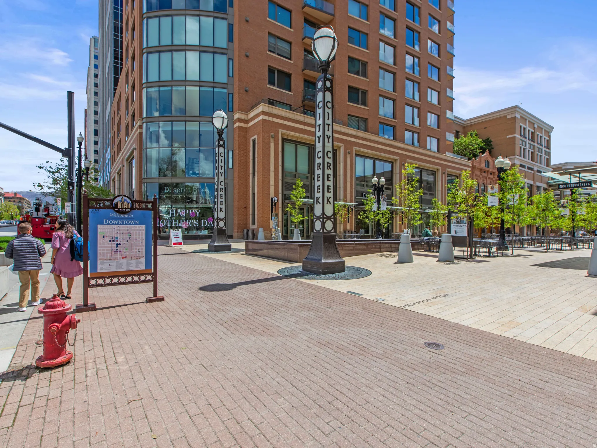 Brick paver walkway with a street lamp featuring "CITY CREEK" signage, ground-level commercial storefronts, and a multi-story brick and glass building